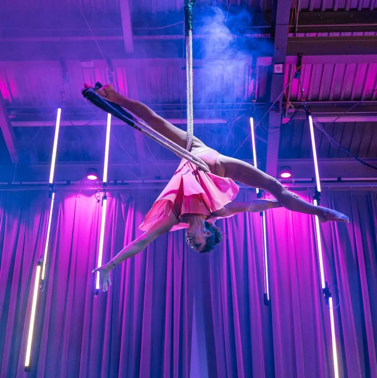 An aerialist in a split on aerial silks under purple light